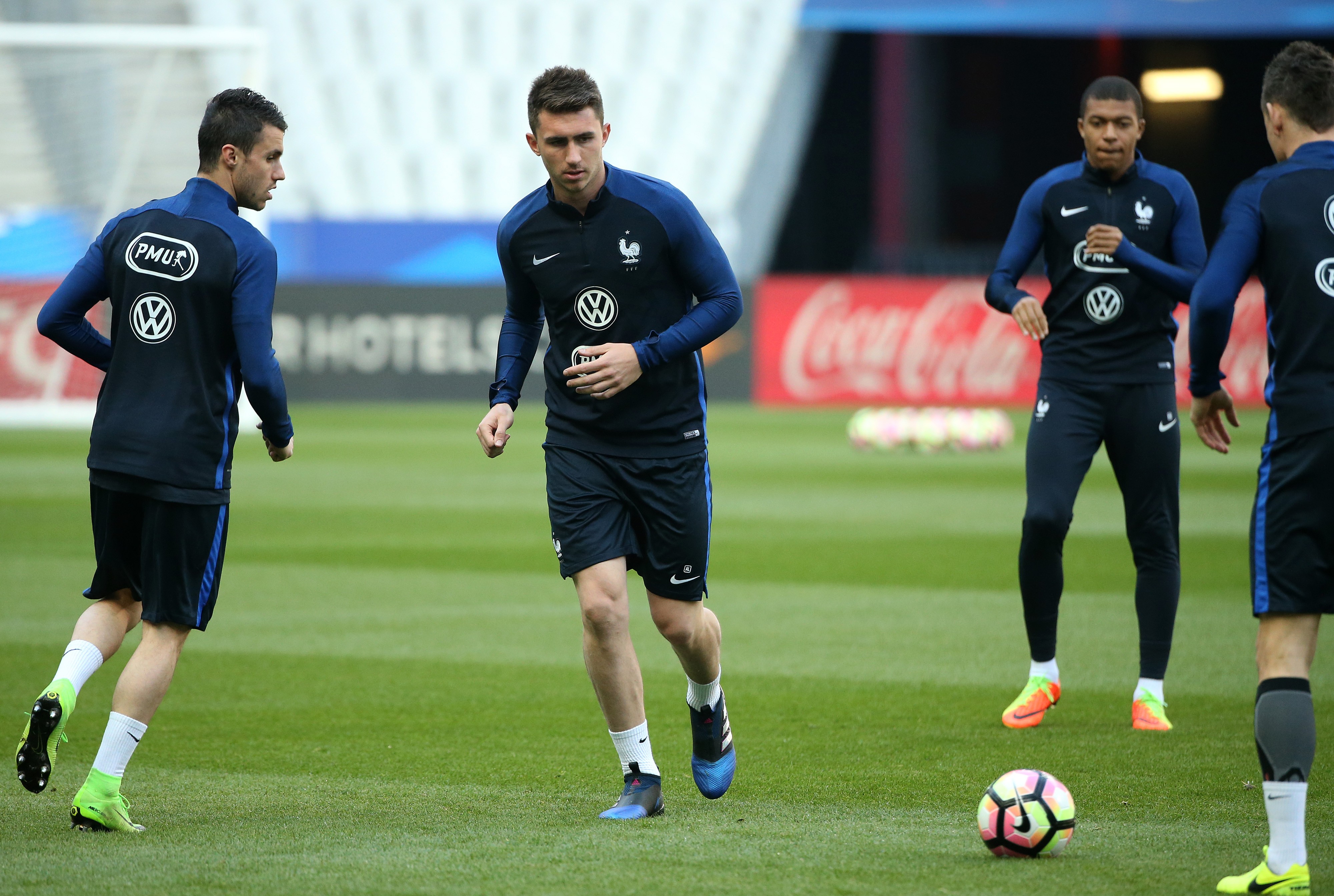 Quatro jogadores de futebol, vestindo uniformes azuis escuros com o logo da Volkswagen, correm em um campo de grama verde. Um jogador no centro olha para frente, enquanto outro à esquerda olha para o lado. Ao fundo, um banner vermelho com o logo da Coca-Cola é visível. Uma bola de futebol colorida está no chão, à direita