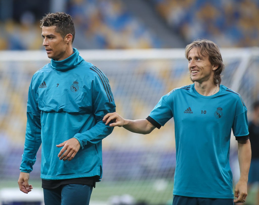 Cristiano Ronaldo, à esquerda, com agasalho azul-petróleo de gola alta, e Luka Modric, à direita, com camisa azul-petróleo de manga curta, sorrindo e com a mão no ombro de Ronaldo, em um campo de futebol