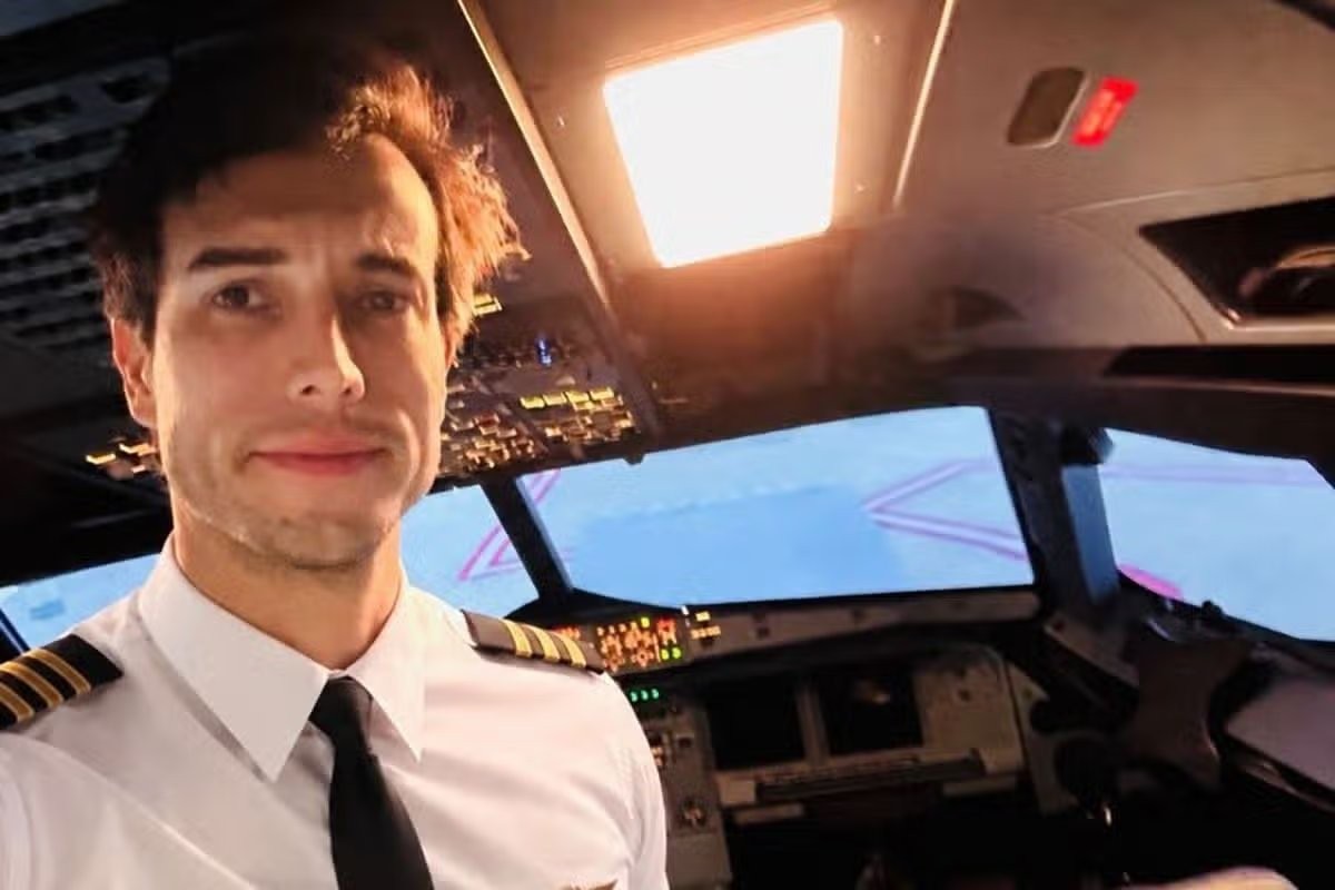 Jovem piloto de avião, com uniforme branco e gravata preta, sorri levemente para a câmera dentro da cabine de comando, com painéis de controle e janelas mostrando uma pista de pouso ao fundo