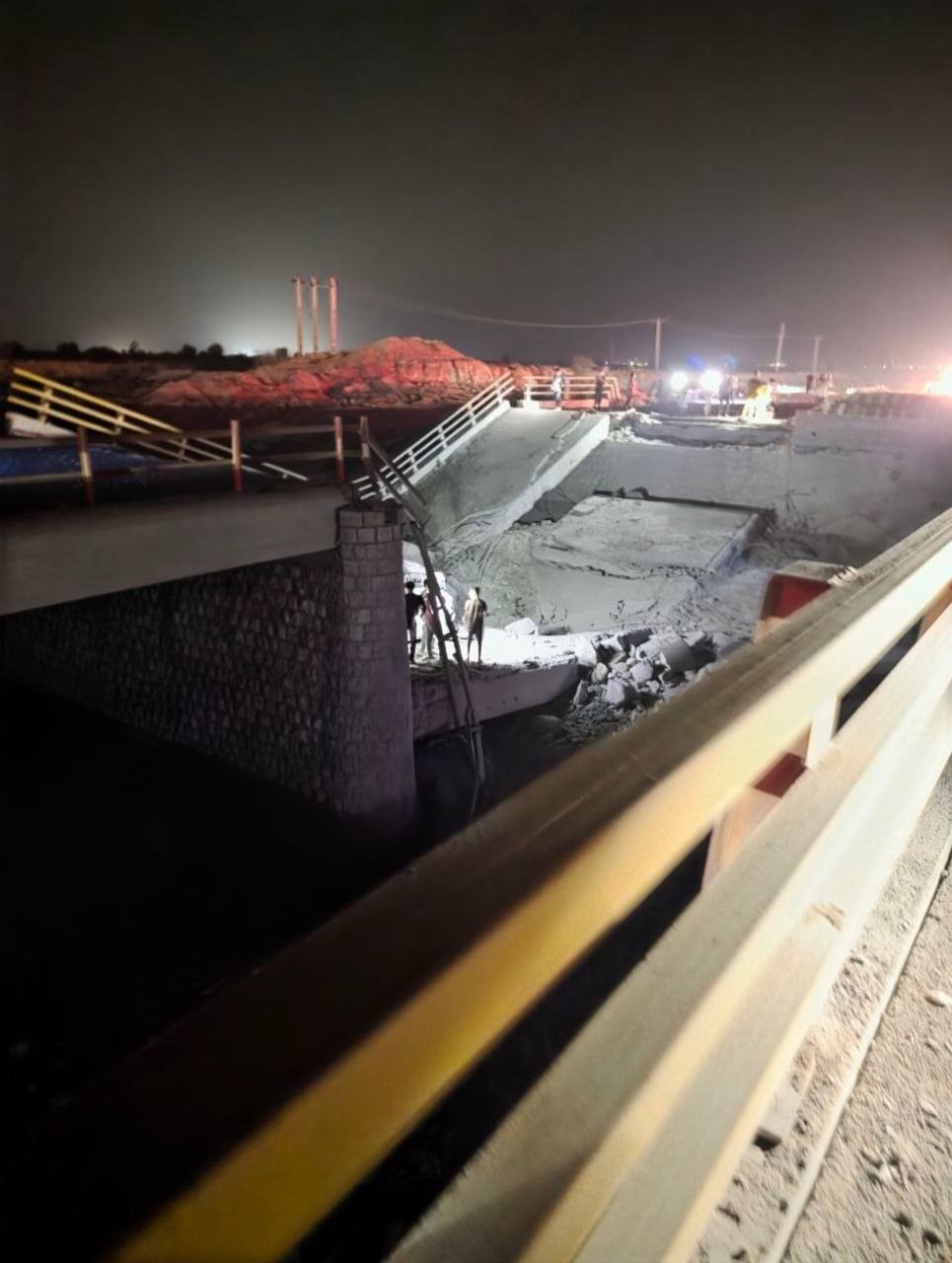 Ponte desabada à noite, com um trecho da estrutura caído no leito seco de um rio. Pessoas com coletes refletivos e capacetes inspecionam os danos, iluminadas por luzes de veículos de emergência. Ao fundo, uma colina avermelhada e o céu escuro.