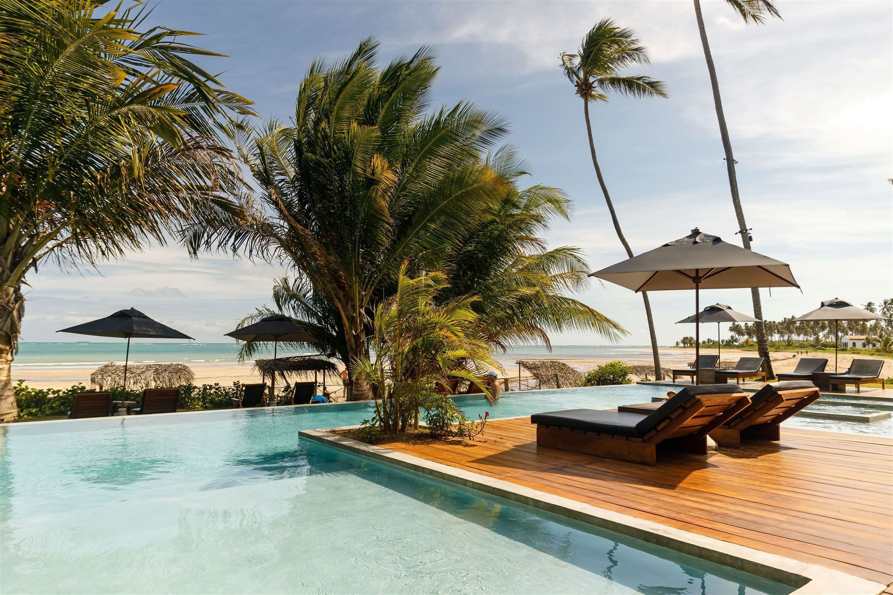 Piscina de resort com borda infinita e deck de madeira, espreguiçadeiras e guarda-sóis. Ao fundo, coqueiros e uma praia de areia clara com o mar azul-turquesa sob céu parcialmente nublado