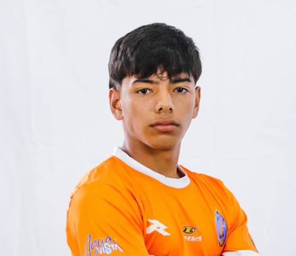 Jovem com pele morena e cabelo escuro, vestindo uma camisa de futebol laranja com gola branca, olhando diretamente para frente com expressão séria