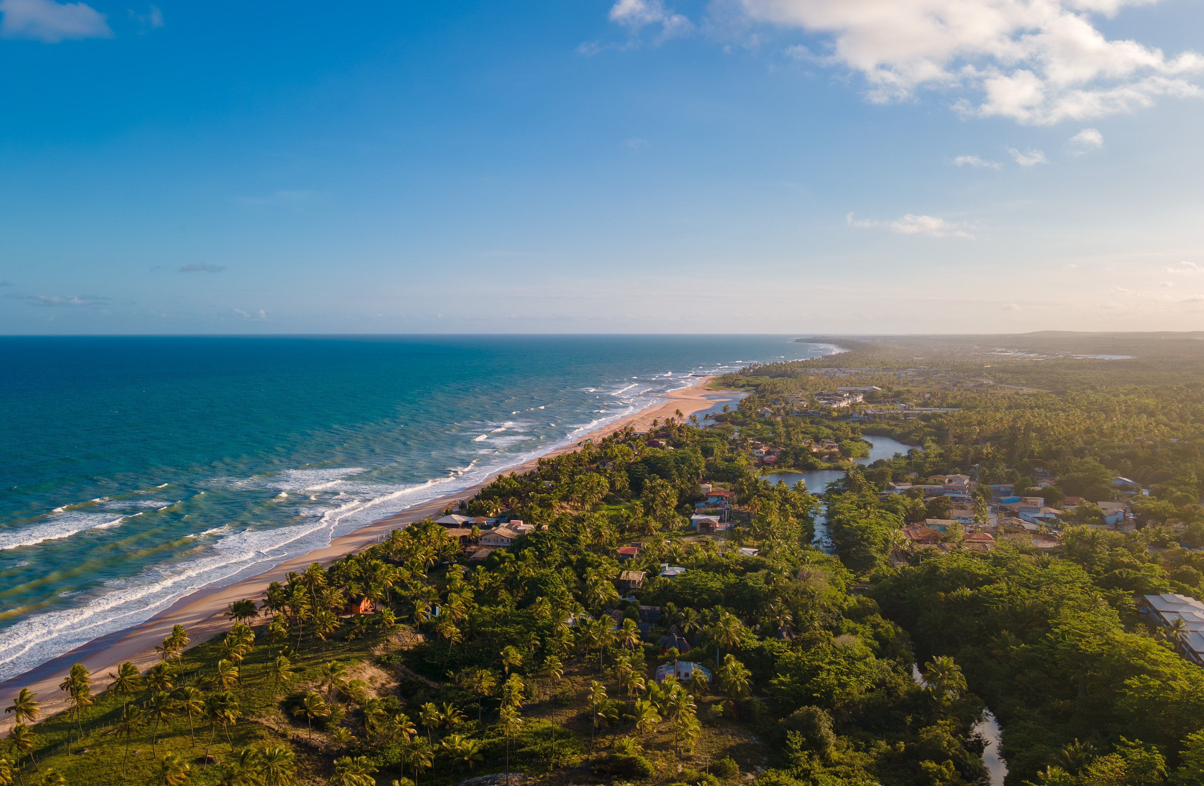 Vista aérea de uma longa praia com areia clara e coqueiros, banhada por um mar azul-turquesa com ondas brancas. À direita, uma área verde densa com casas e um rio sinuoso, sob um céu azul com nuvens esparsas e luz dourada do sol poente