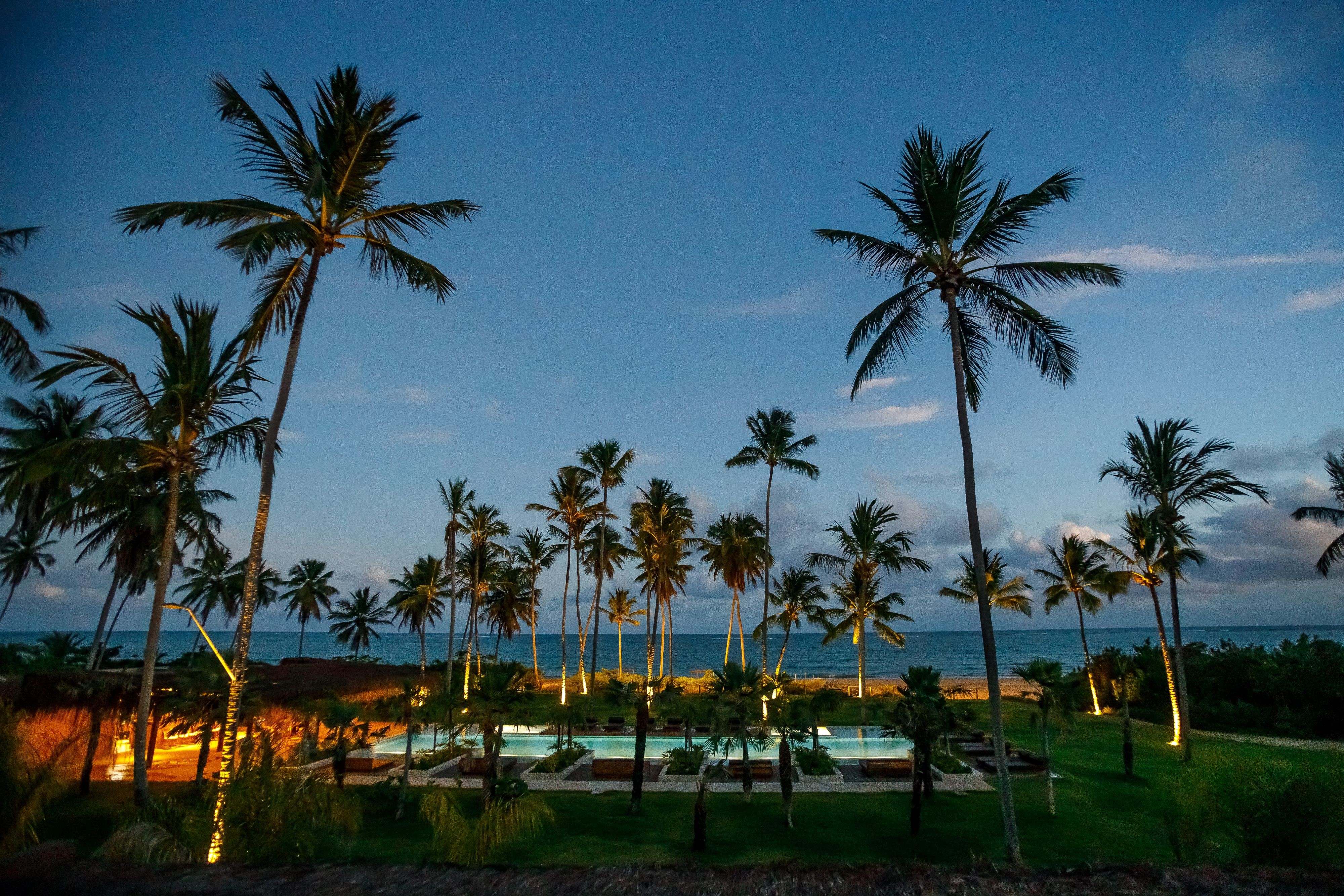 Cenário noturno de um resort tropical com coqueiros altos iluminados, piscinas azuis e o mar ao fundo sob um céu escuro com nuvens claras