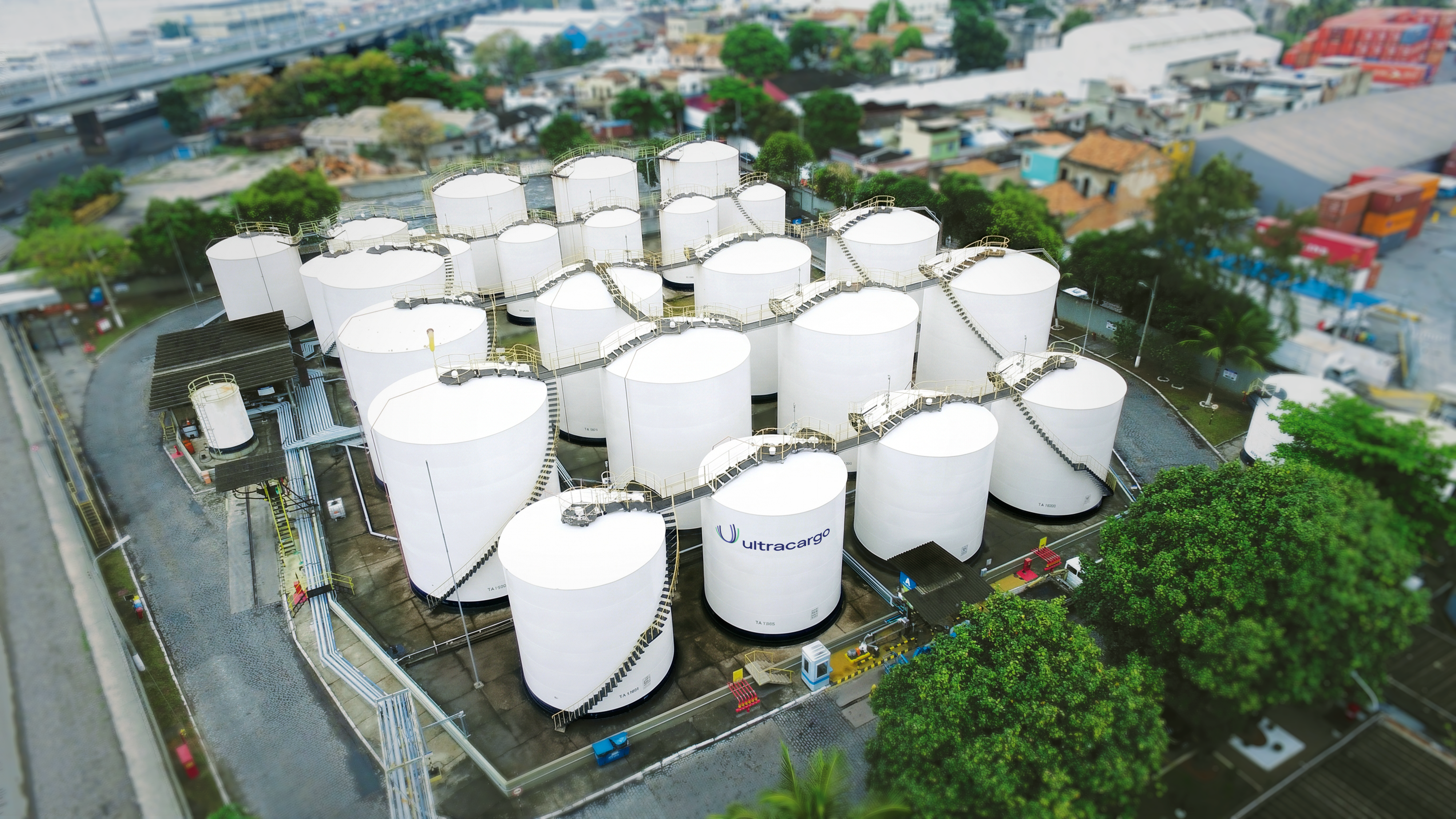 Vista aérea de um complexo industrial com dezenas de grandes tanques brancos cilíndricos, alguns com o logo ultracargo, interligados por tubulações e passarelas. Ao fundo, uma ponte, áreas urbanas e vegetação.