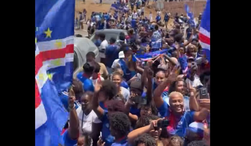 Multidão de pessoas, muitas vestindo azul, celebrando e acenando em uma rua, com bandeiras de Cabo Verde visíveis
