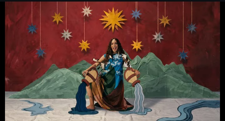 Mulher de cabelos longos e escuros, vestindo um vestido colorido, sentada com as pernas abertas, segurando dois jarros que derramam água azul. Ao fundo, um cenário pintado com montanhas verdes e um céu vermelho com estrelas penduradas
