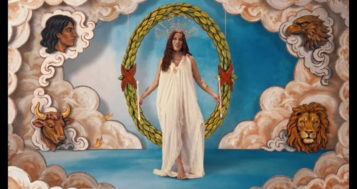 Mulher de cabelos longos e coroa dourada, vestindo um longo vestido branco, em pé no centro de uma coroa de louros gigante. Ao redor, nuvens com cabeças de homem, touro, águia e leão, sobre fundo azul
