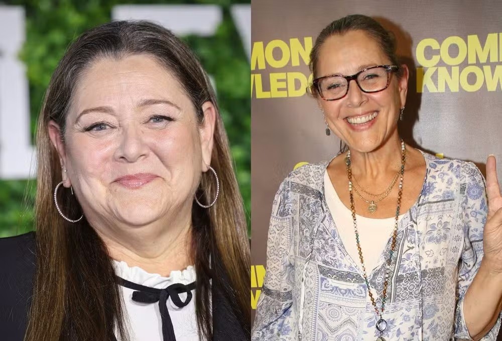 Duas mulheres sorridentes, a da esquerda com cabelos castanhos longos e brincos de argola, a da direita com óculos e colar, fazendo sinal de paz