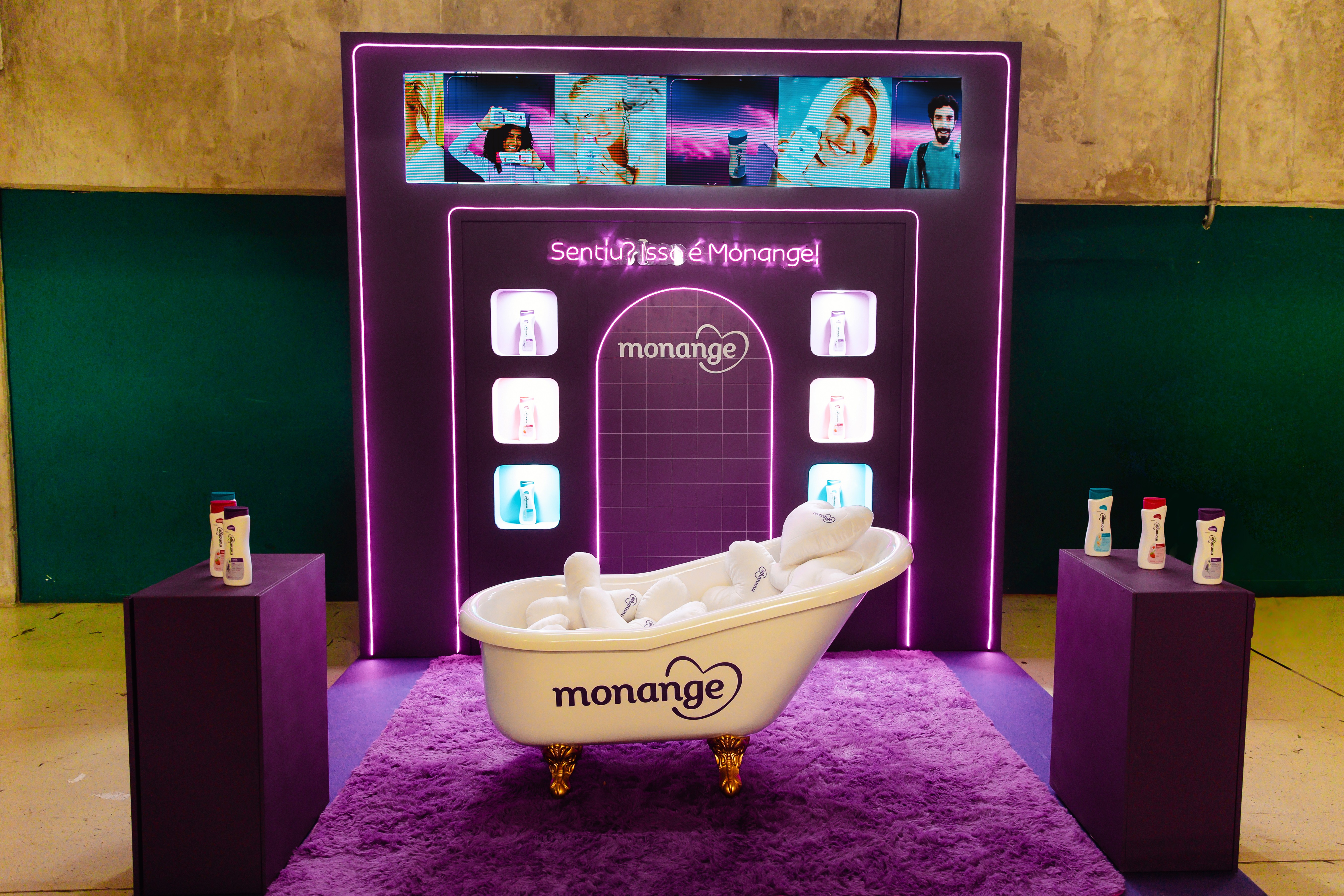 Cen&aacute;rio de evento Monange com banheira branca cheia de toalhas, tapete roxo felpudo, painel neon roxo com a frase Sentir isso &eacute; Monange e telas exibindo pessoas felizes. Dois pedestais roxos exibem produtos da marca