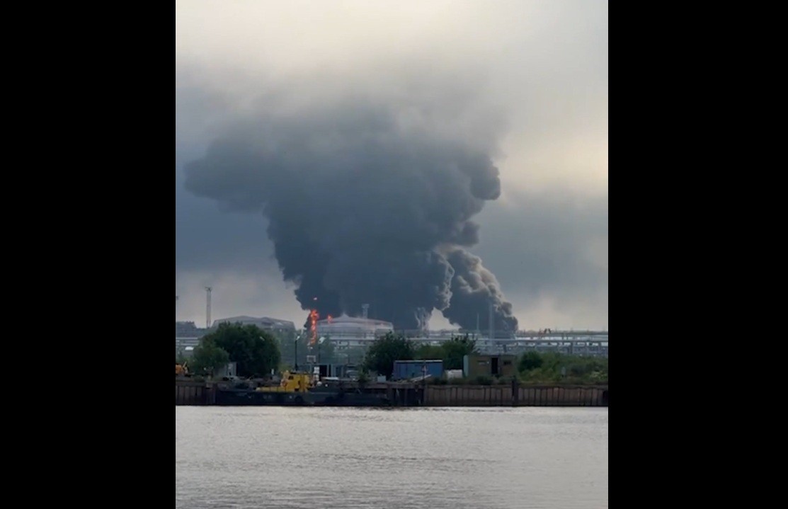 Fumaça escura e densa sobe de um incêndio em uma instalação industrial à beira de um rio, com chamas visíveis em um dos edifícios