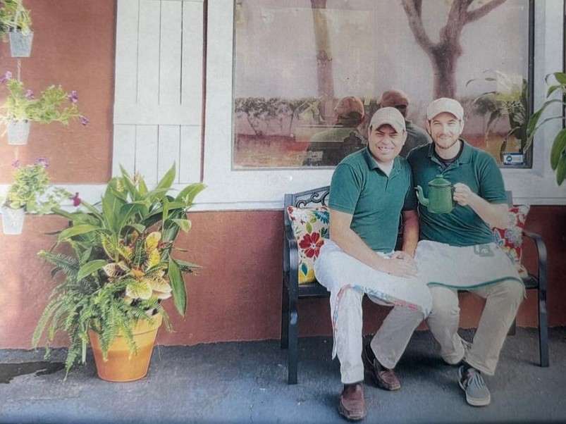 Dois homens sorridentes, de boné e camisa polo verde, sentados em um banco de ferro com almofadas florais. O da direita segura um bule verde. Ao fundo, uma janela reflete árvores e pessoas, e à esquerda, plantas em vasos decoram a parede.
