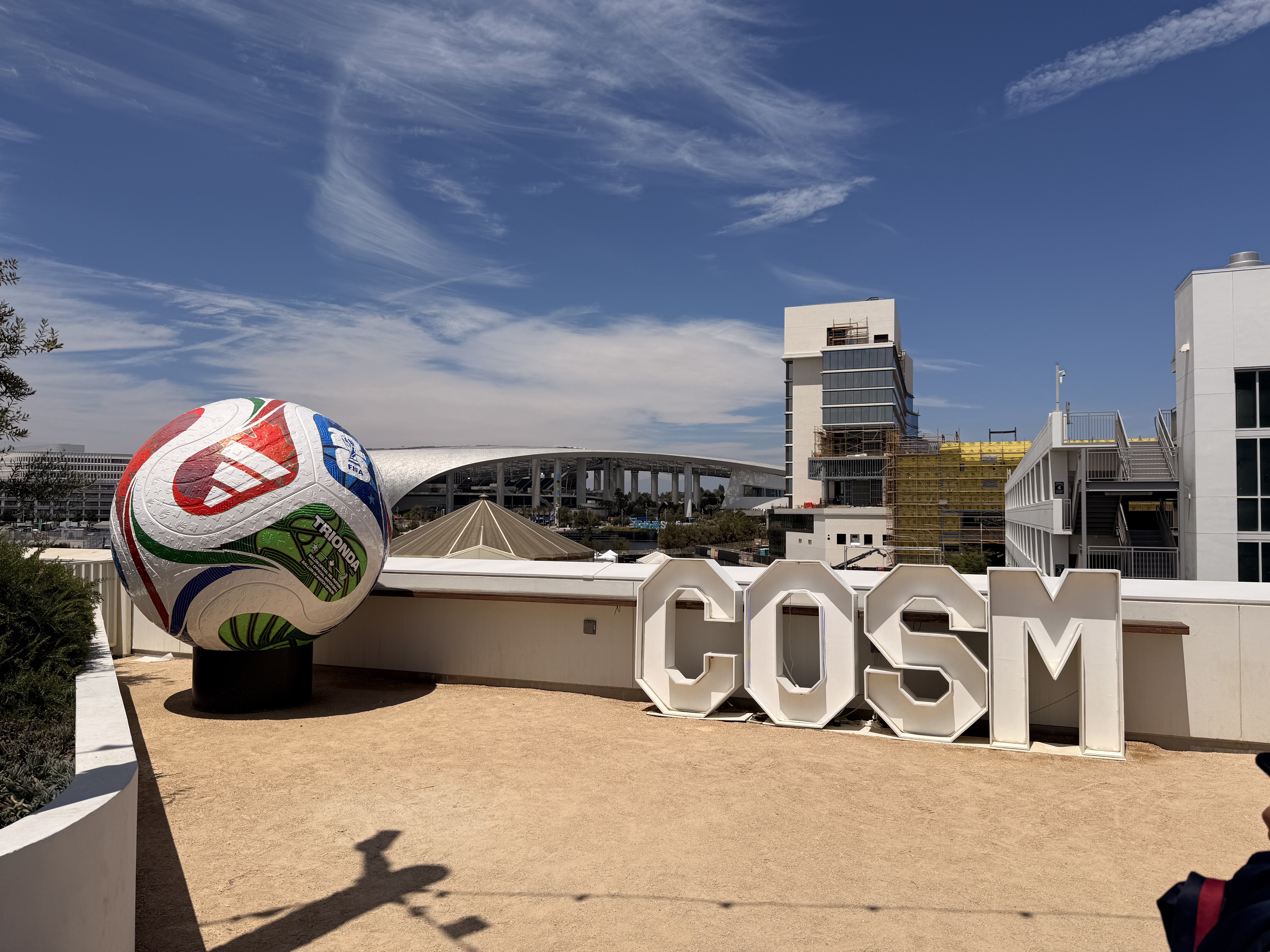 Bola de futebol gigante colorida com o nome TRIOMDA e o logotipo da Adidas, ao lado das letras brancas COSM em um terraço. Ao fundo, um estádio moderno com teto curvo e edifícios altos sob céu azul com nuvens esparsas