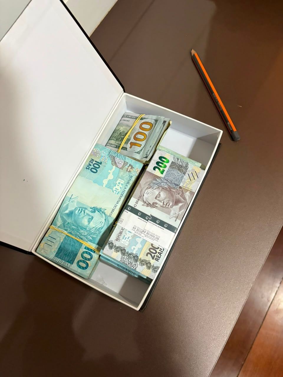 Caixa branca aberta com ma&ccedil;os de dinheiro: um de notas de 100 d&oacute;lares, um de 100 reais e outro de 200 reais. Um l&aacute;pis est&aacute; ao lado da caixa sobre uma superf&iacute;cie marrom escura