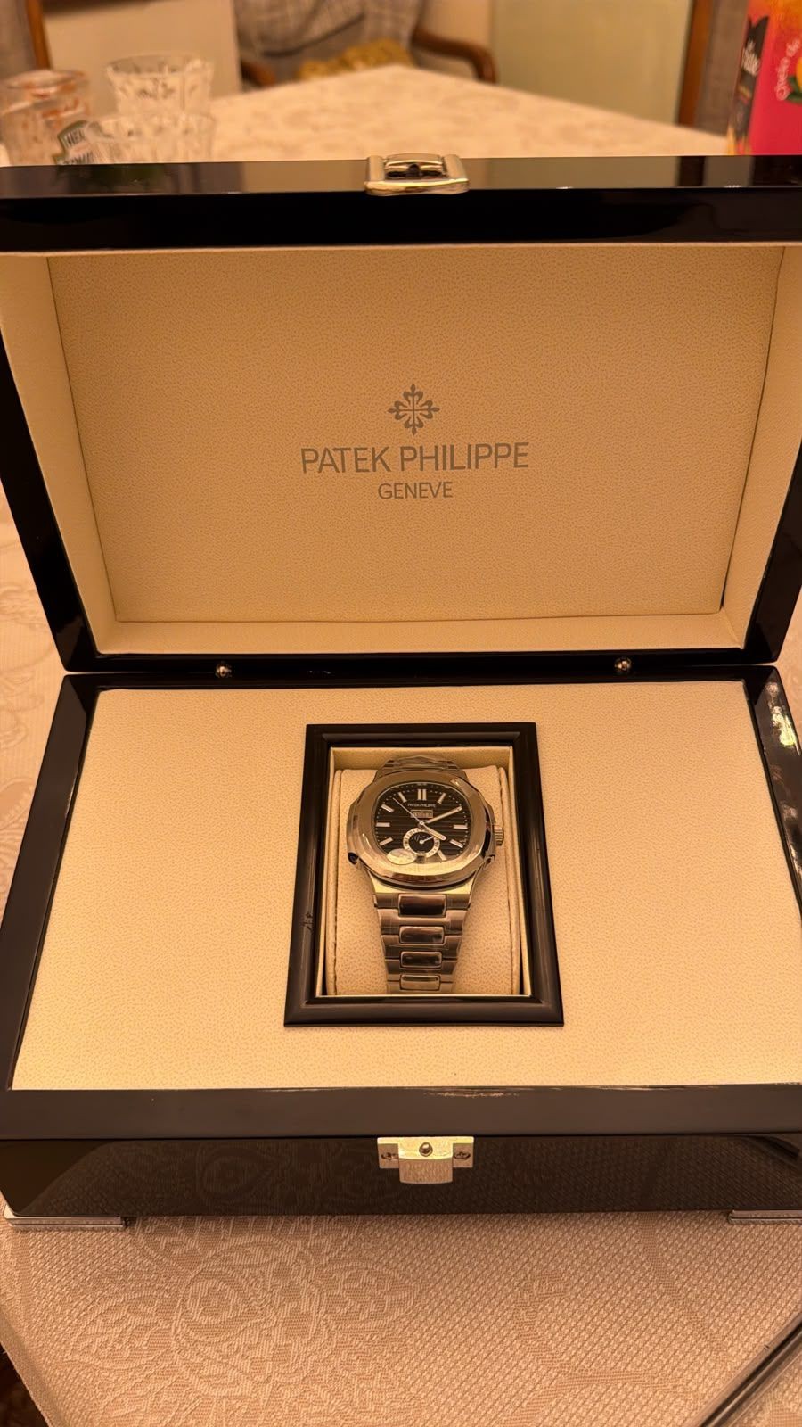 Rel&oacute;gio Patek Philippe Nautilus de a&ccedil;o inoxid&aacute;vel com mostrador preto, dentro de uma caixa preta aberta com interior bege, sobre uma toalha de mesa clara
