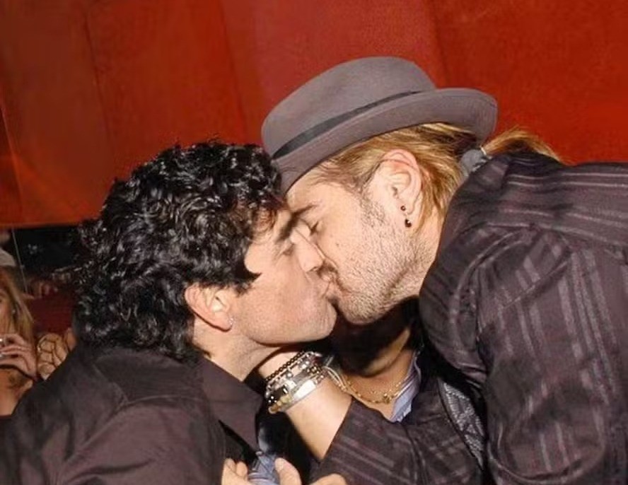 Dois homens se beijando intensamente. Um tem cabelo escuro e encaracolado, camisa preta. O outro, de barba, cabelo loiro preso e chapéu cinza, usa camisa listrada e pulseiras. Fundo vermelho borrado