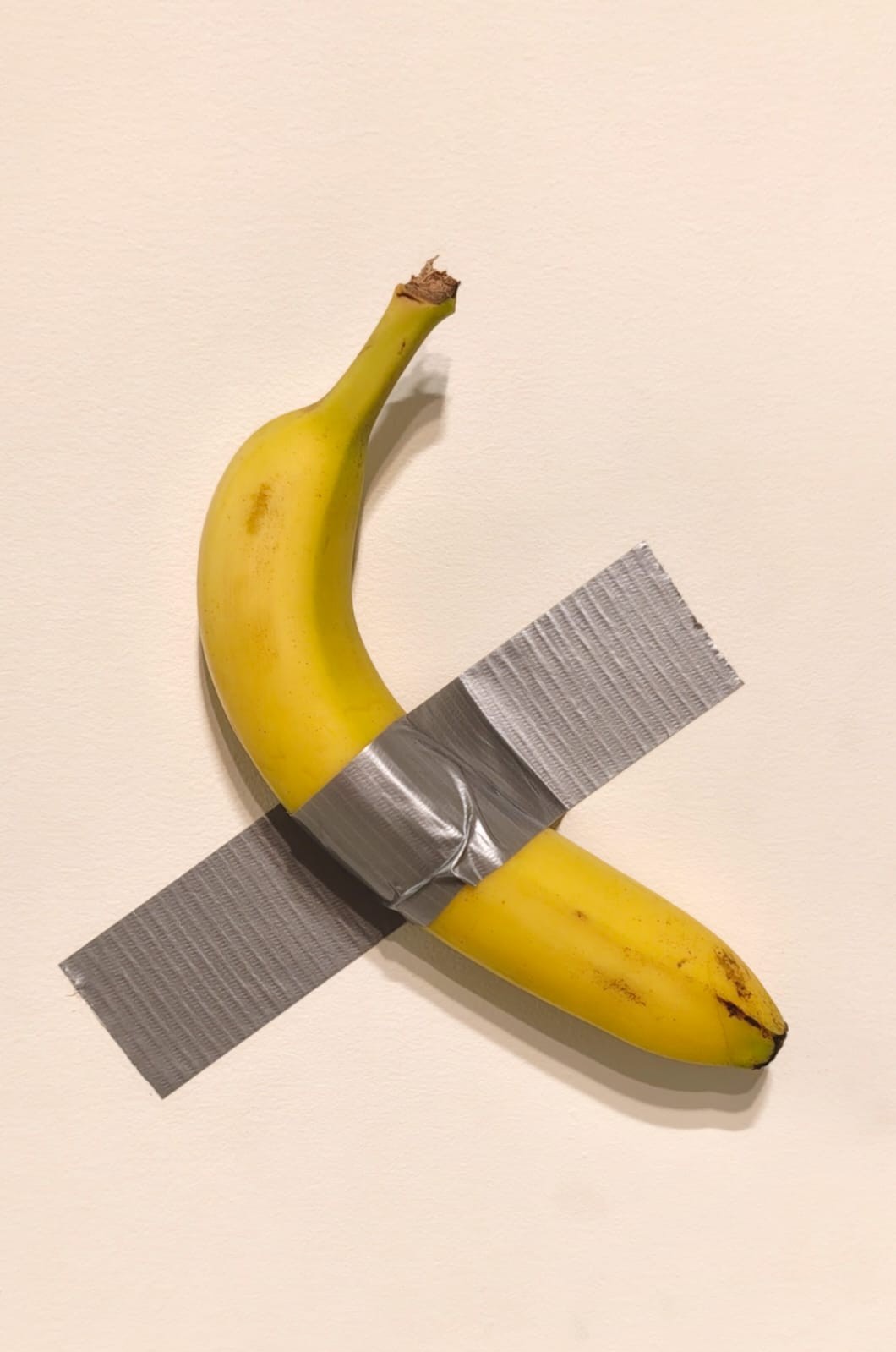Banana madura amarela presa verticalmente a uma parede clara com duas tiras de fita adesiva cinza, formando um X
