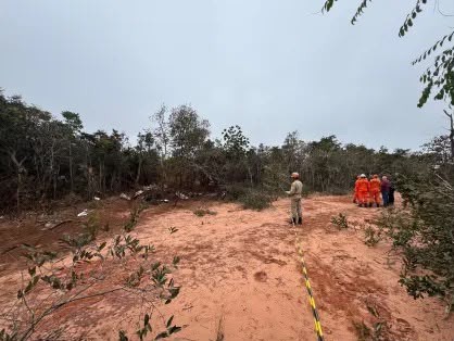 Equipe de resgate em área de mata com solo avermelhado e vegetação rasteira, sob céu nublado. Um homem de uniforme claro está de costas, e um grupo de pessoas com uniformes laranja está à direita, próximos à mata. Uma fita de isolamento amarela e preta demarca o chão.