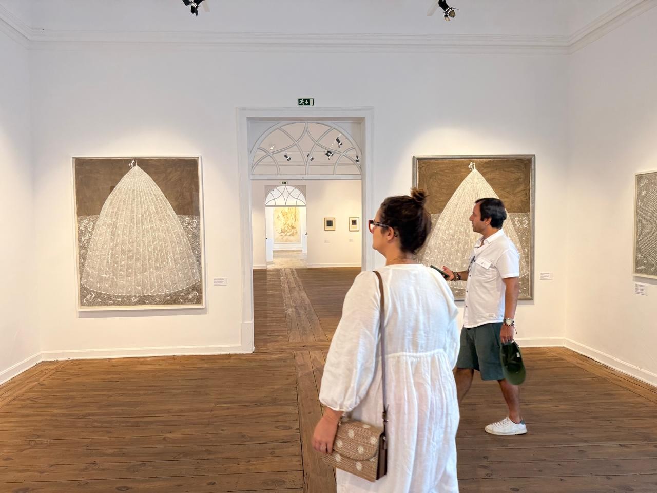 Uma mulher de costas, com vestido branco e bolsa bege de bolinhas, observa pinturas em uma galeria de arte. Ao lado, um homem de camisa branca e bermuda verde também admira as obras. O ambiente tem paredes brancas, piso de madeira e quadros com formas triangulares brancas sobre fundo marrom. Ao fundo, um corredor com mais obras e uma porta arqueada.