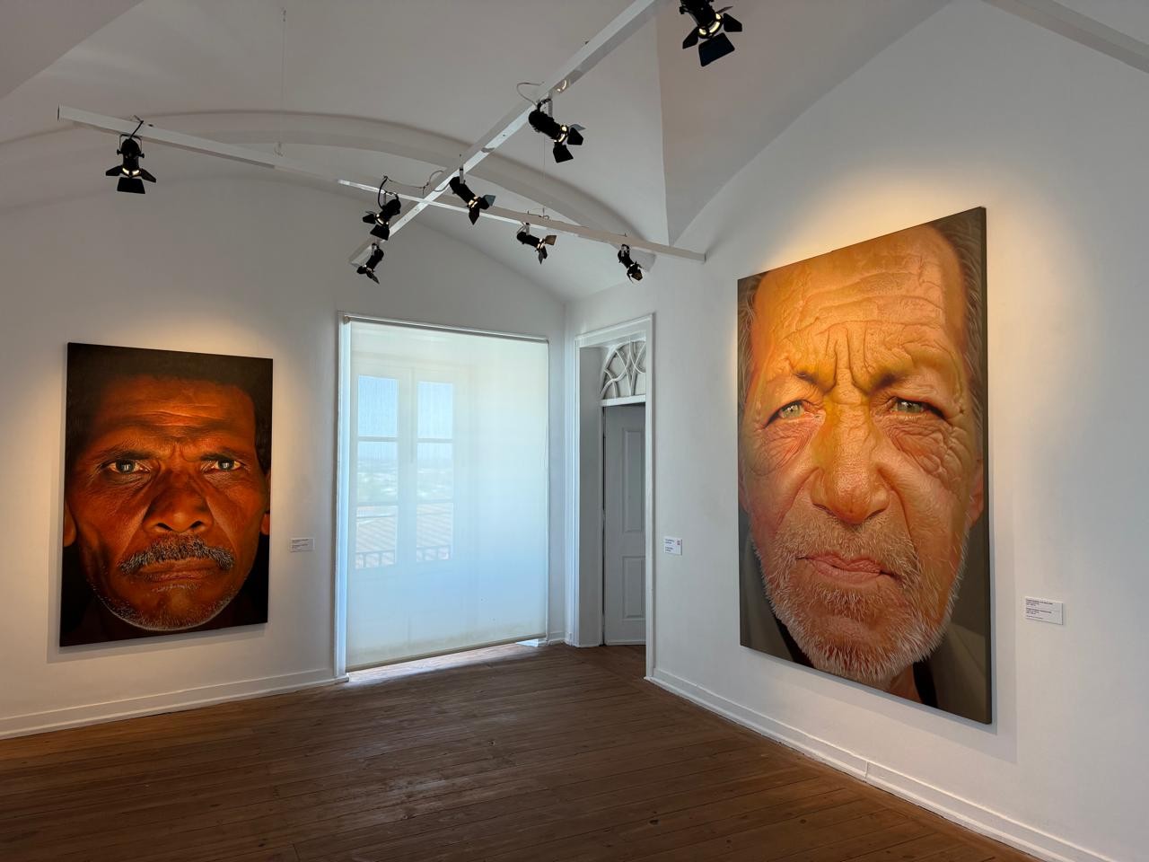 Galeria de arte com paredes brancas e piso de madeira, exibindo dois grandes retratos de homens. À esquerda, um homem de pele escura com bigode e olhar sério. À direita, um homem branco de olhos azuis, barba grisalha e expressão franzida. Focos de luz no teto iluminam as obras
