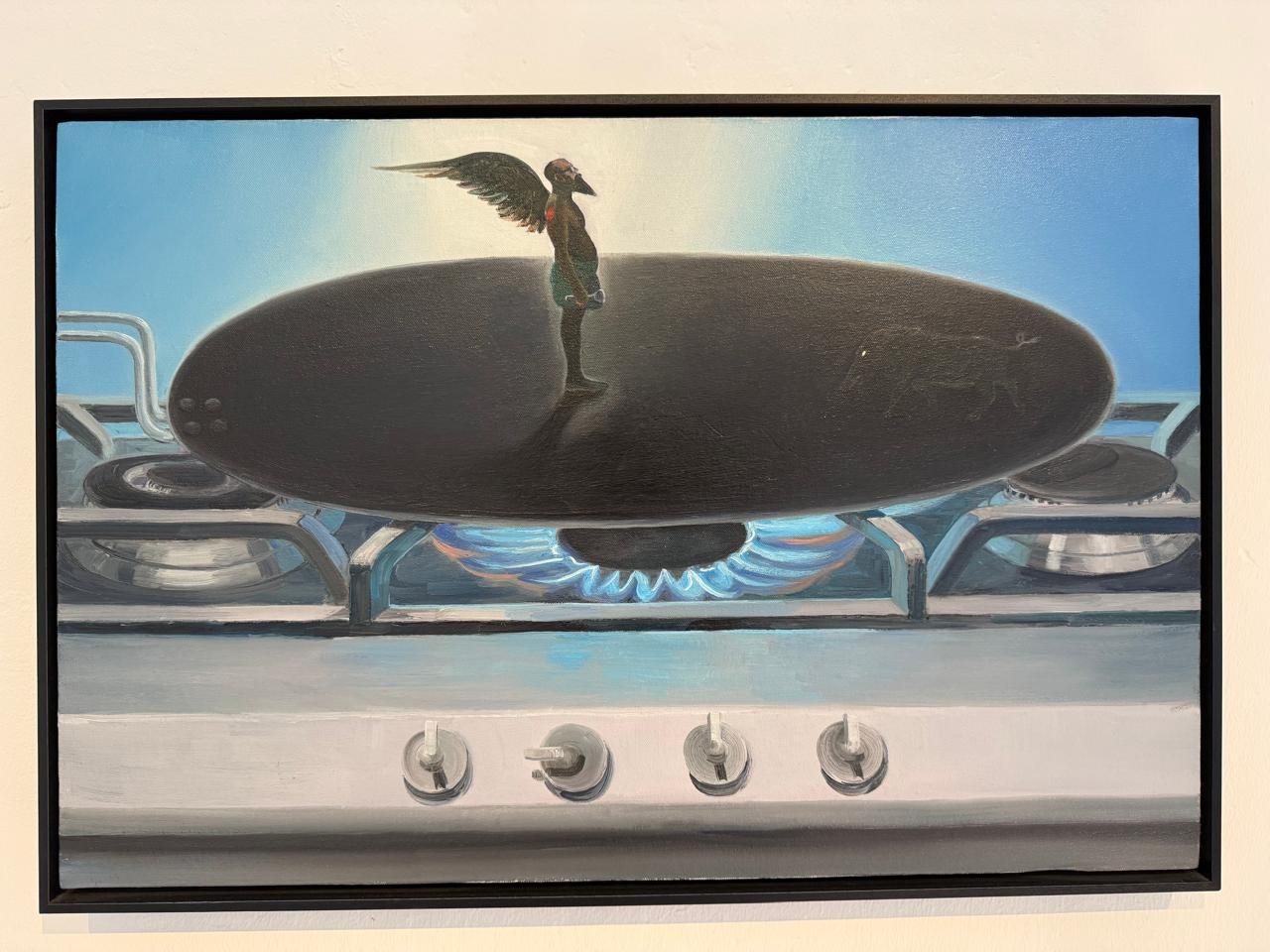 Pintura surrealista de um fogão branco com chamas azuis acesas, sobre o qual repousa uma frigideira oval escura. No centro da frigideira, uma figura humana alada, com cabeça de pássaro e corpo esguio, está de pé, olhando para a direita. O fundo é um céu azul claro com um brilho branco no horizonte. A obra está em uma moldura preta.