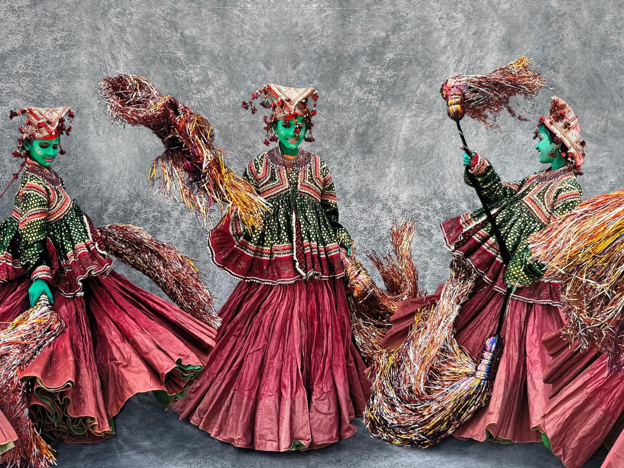 Três pessoas com rostos pintados de verde e chapéus ornamentados, vestindo roupas tradicionais em tons de verde e vermelho, com saias rodadas e longas. Elas seguram vassouras decoradas com fibras coloridas, em um fundo cinza texturizado