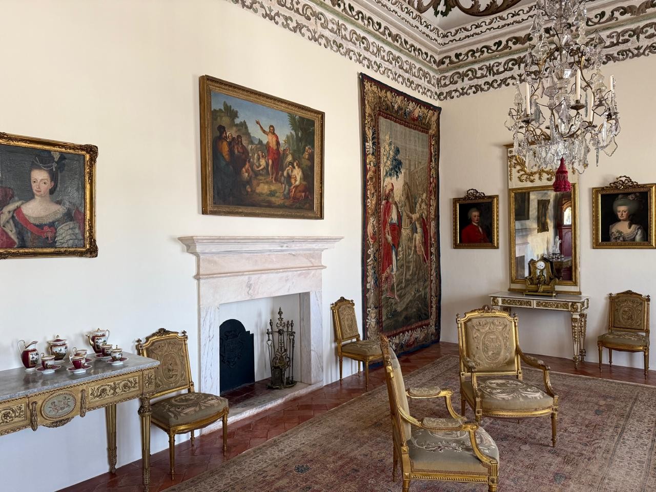 Salão de um palácio com paredes brancas, teto decorado, lareira de mármore, pinturas a óleo, tapeçaria, espelho dourado, lustre de cristal e cadeiras ornamentadas com estofamento claro. Um console com serviço de chá está à esquerda. O chão tem um tapete persa sobre piso de terracota