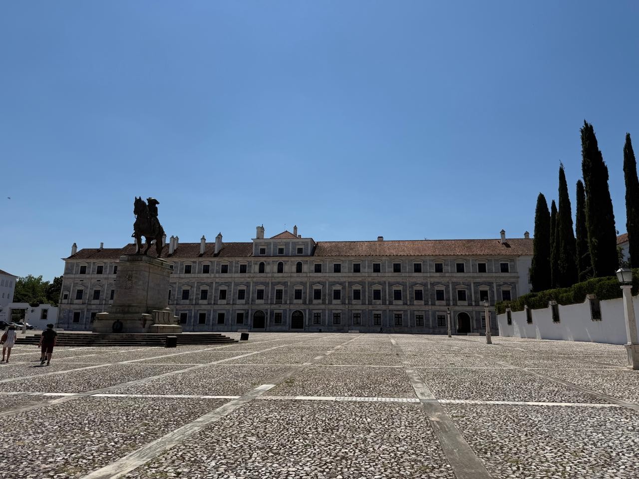 Praça com calçamento de pedras claras e escuras, estátua equestre de bronze à esquerda, e um grande edifício histórico de três andares com telhado de telhas marrons ao fundo. À direita, ciprestes altos e um muro branco. O céu é azul claro e sem nuvens
