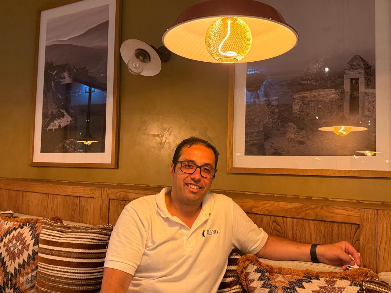 Homem de óculos e camisa polo branca sorri sentado em sofá com almofadas estampadas, sob luminárias e quadros na parede