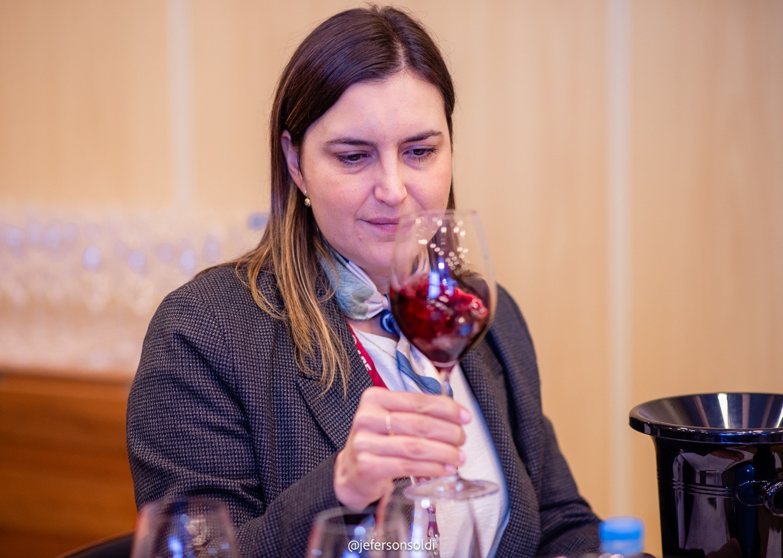 Mulher de cabelos castanhos, vestindo blazer cinza e lenço estampado, cheira um copo de vinho tinto com expressão concentrada, em um ambiente interno com fundo bege