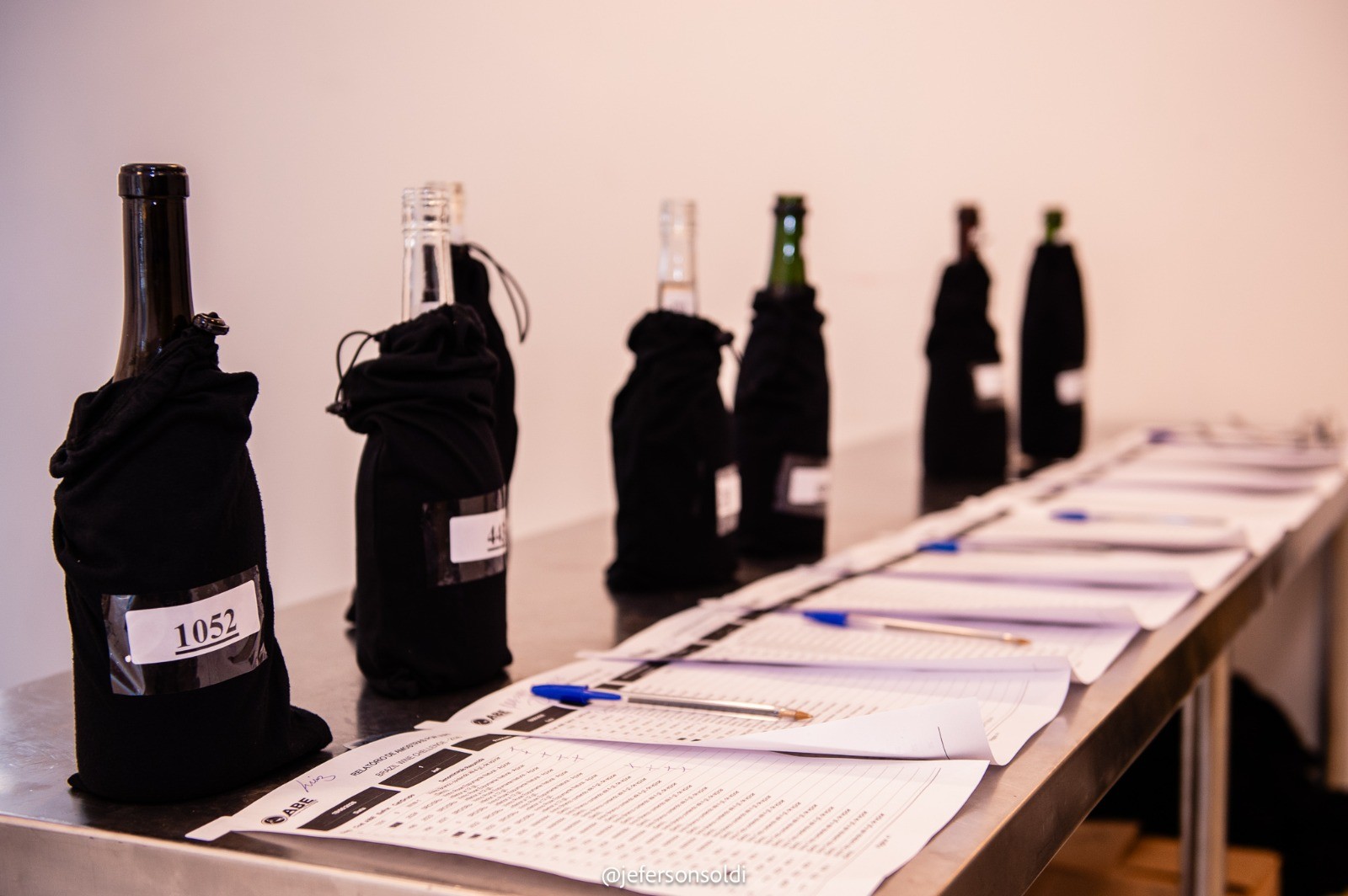 Garrafas de vinho cobertas com sacos pretos e numeradas, alinhadas em uma mesa metálica com formulários e canetas para degustação às cegas