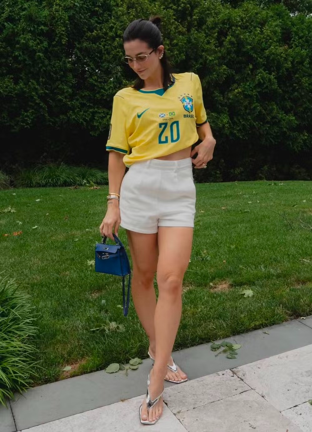 Mulher de óculos, cabelo preso, vestindo camisa amarela da seleção brasileira com o número 20, shorts brancos e chinelos prateados, segurando uma pequena bolsa azul, em pé sobre um caminho de pedras com grama e arbustos verdes ao fundo