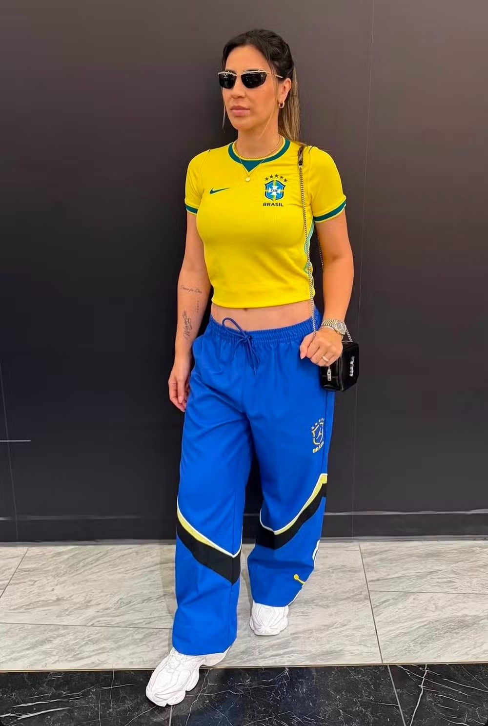 Mulher de óculos escuros, camiseta amarela da seleção brasileira e calça azul com detalhes amarelos e pretos, posando em pé contra uma parede escura e piso de mármore
