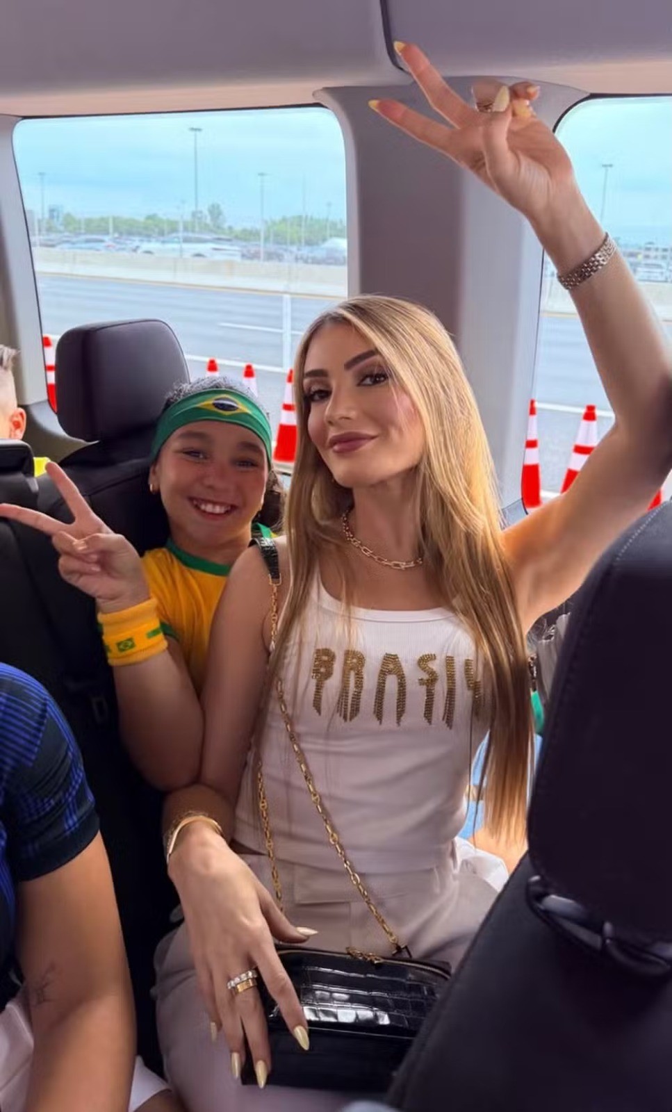 Mulher loira sorridente e criança com bandana do Brasil fazem sinal de paz dentro de um veículo, com cones de trânsito visíveis pela janela