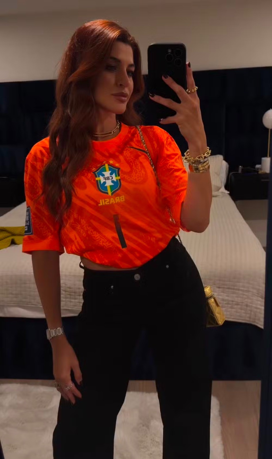 Mulher de cabelos castanhos avermelhados, usando camisa laranja da seleção brasileira, calça preta e joias douradas, tira selfie no espelho de um quarto