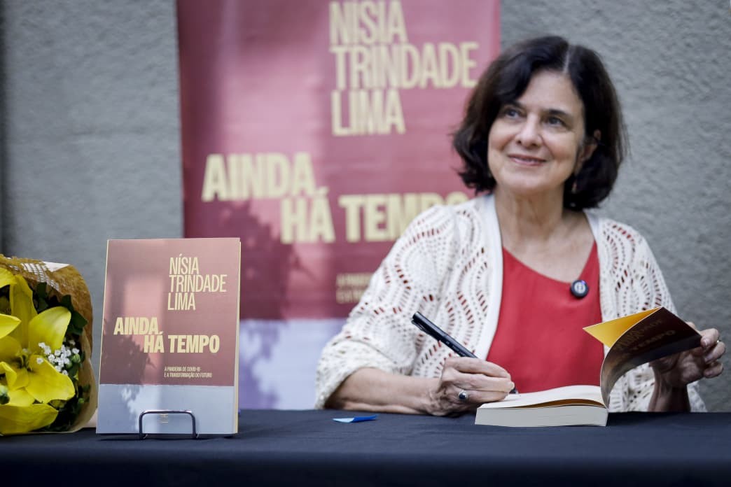 Nísia Trindade Lima, com cabelo escuro e ondulado, sorri enquanto autografa um livro. Ela veste uma blusa vermelha e um cardigã de tricô branco, sentada a uma mesa preta. À esquerda, um buquê de flores amarelas e um livro em um suporte, com o título Ainda Há Tempo. Ao fundo, um banner vermelho com o nome dela e o mesmo título