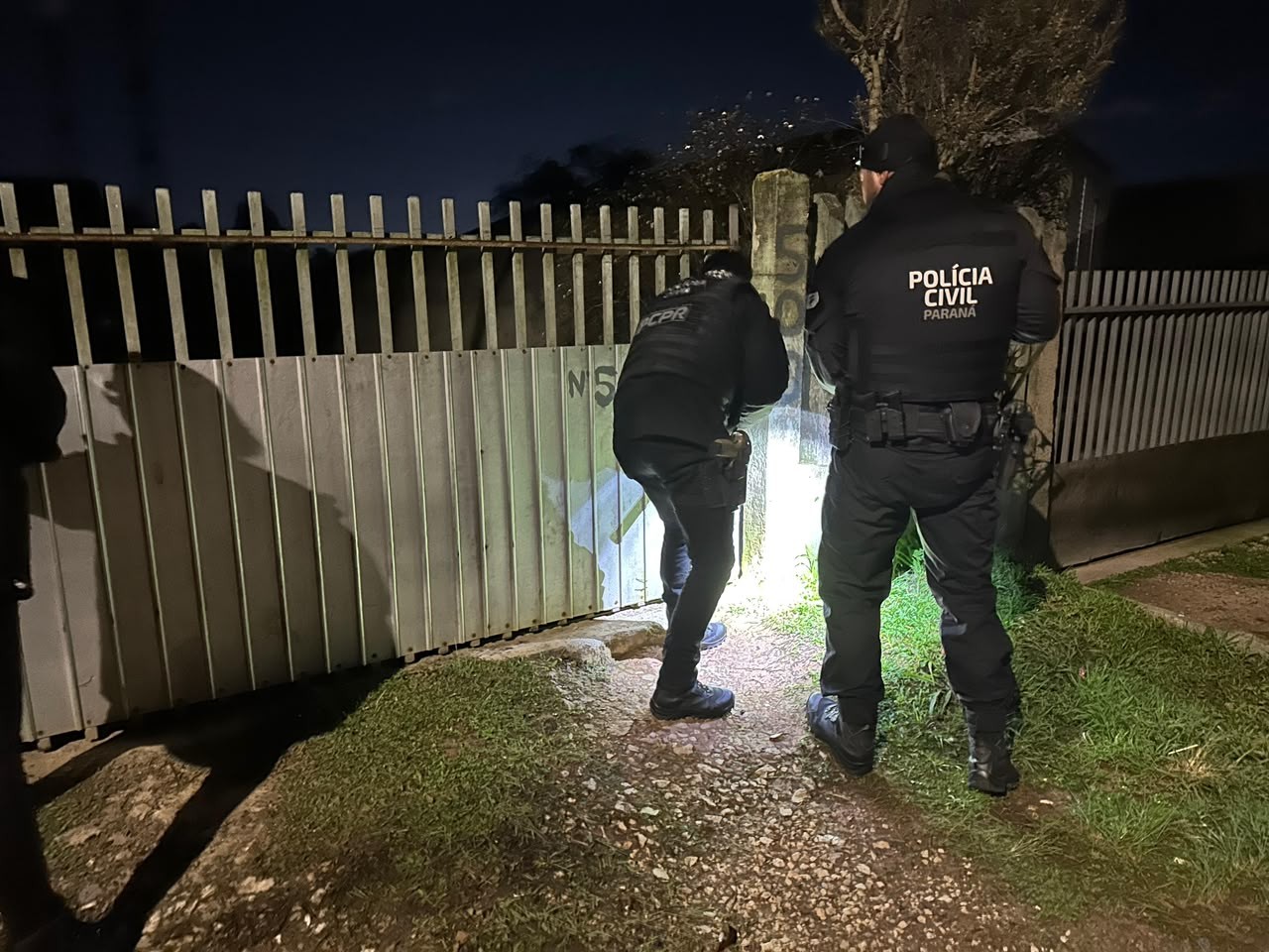 Dois policiais civis do Paraná, vestidos de preto, inspecionam um portão metálico cinza à noite, um deles agachado com uma lanterna, iluminando o chão e a base do portão. O outro policial está de costas, com a inscrição POLÍCIA CIVIL PARANÁ visível em sua jaqueta. O cenário é escuro, com grama e terra no chão