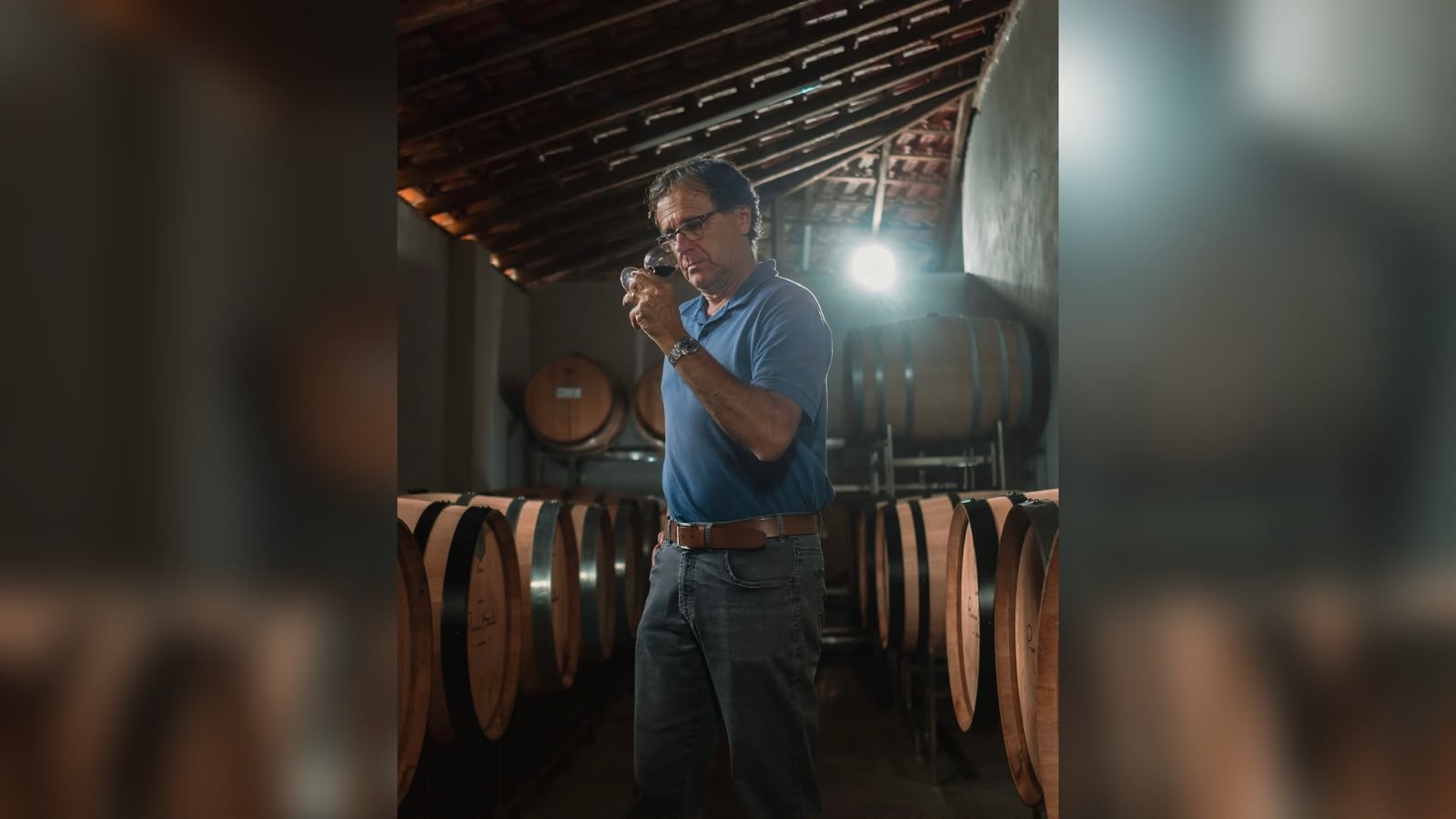 Homem de óculos, camisa polo azul e calça jeans, cheira um copo de vinho tinto em uma adega escura, cercado por barris de madeira