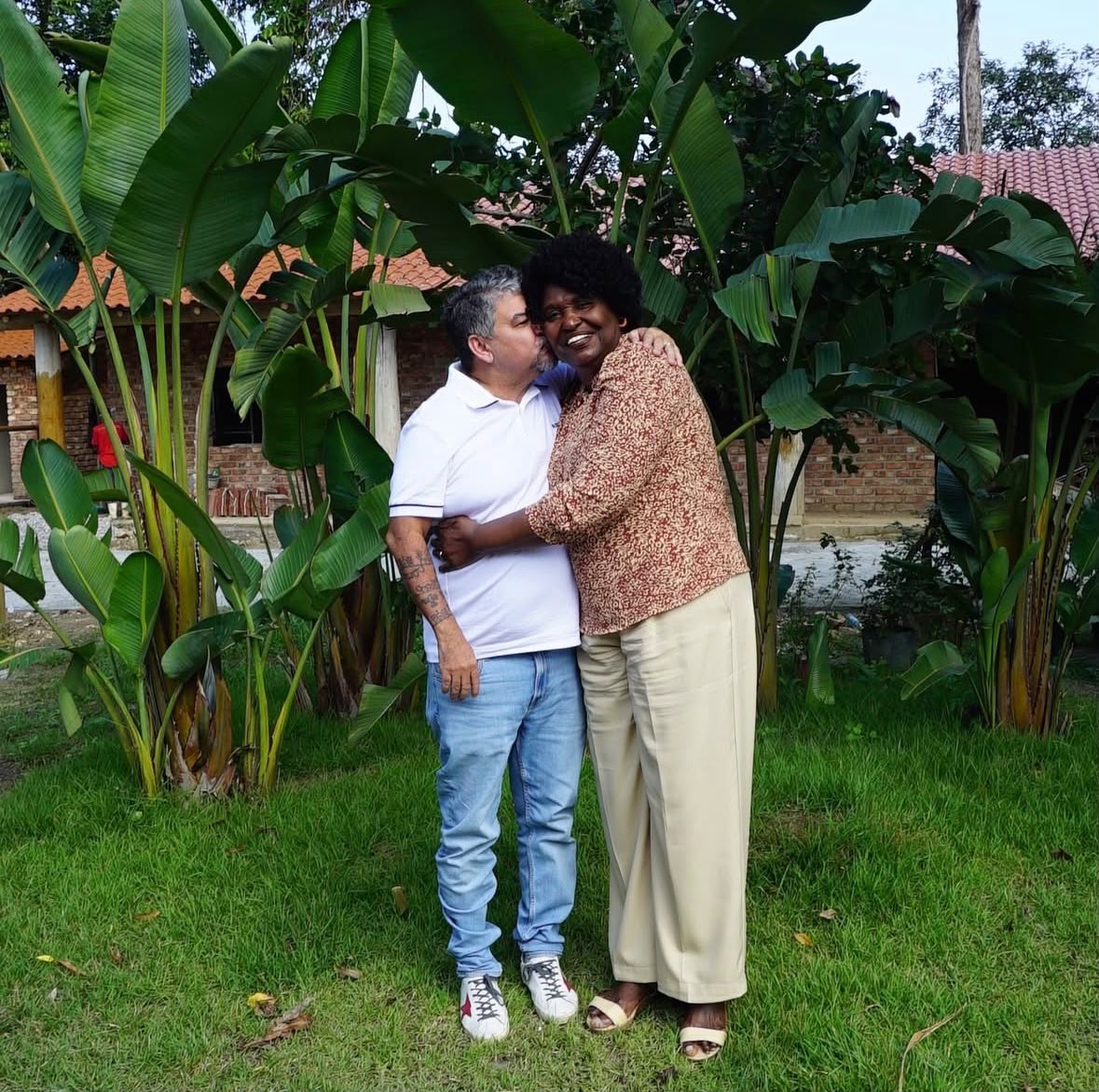 Um homem de pele clara, cabelo grisalho e camiseta branca beija a bochecha de uma mulher negra de cabelo crespo e blusa estampada, ambos sorrindo e abraçados em um jardim com plantas grandes e uma casa ao fundo