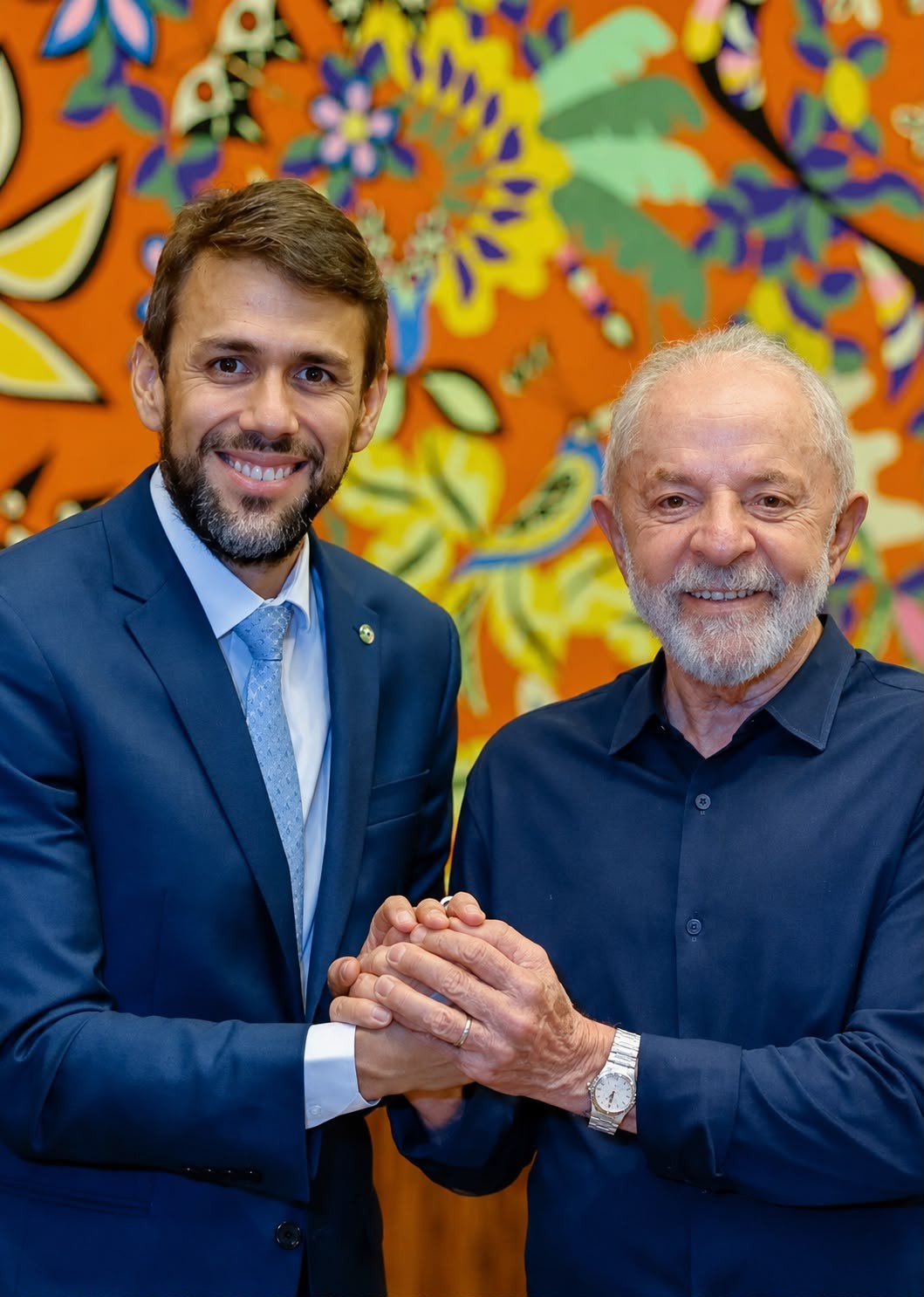Dois homens sorridentes, um jovem de terno azul e outro mais velho de camisa azul-marinho, apertam as mãos em frente a um painel colorido com motivos florais