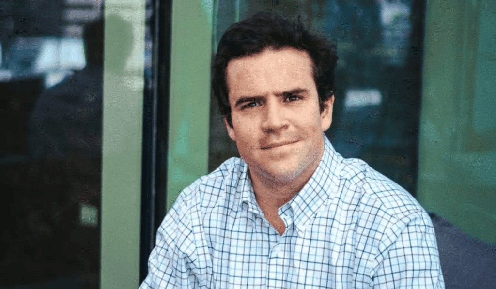 Homem branco de cabelo escuro e camisa xadrez azul e branca, sorrindo levemente, com fundo de vidro verde e reflexos