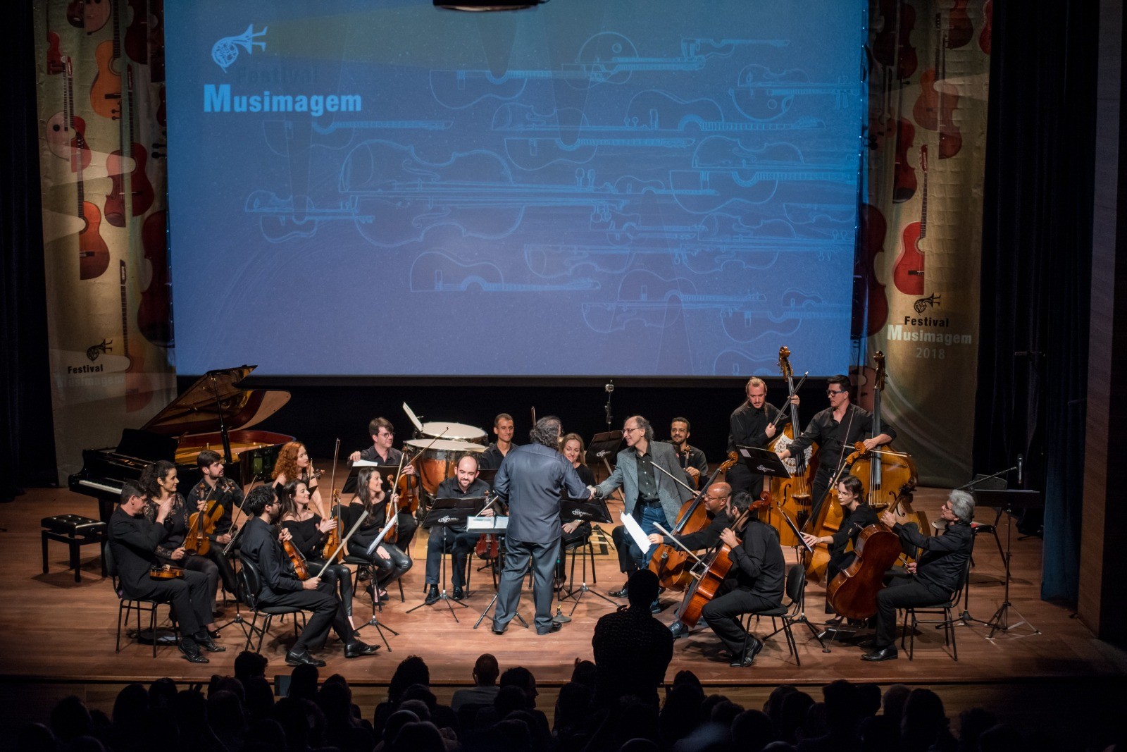 Orquestra com cerca de 20 músicos tocando instrumentos de cordas, percussão e piano em um palco de madeira, com um telão azul ao fundo exibindo o nome Musimagem e um banner vertical com desenhos de violinos