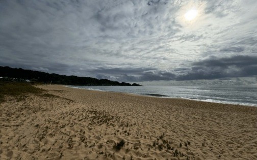 Praia de areia clara com pequenas ondulações, mar azul-escuro com ondas suaves e uma faixa de vegetação rasteira à esquerda. Ao fundo, morros cobertos por mata densa sob um céu nublado com o sol brilhando intensamente no canto superior direito