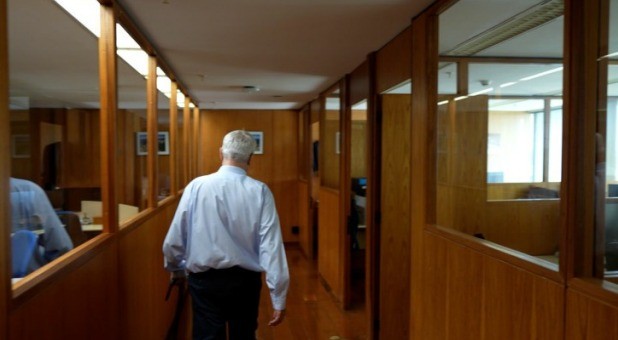 Homem idoso de cabelos brancos, camisa social azul clara e calça escura, visto de costas, caminhando por um corredor de escritório com paredes de madeira e divisórias de vidro, refletindo a iluminação interna