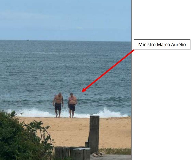 Dois homens de meia-idade, um deles identificado como Ministro Marco Aurélio, saem do mar em uma praia de areia clara, com ondas pequenas e céu nublado. Ambos usam apenas shorts de banho escuros, com o homem à direita, o ministro, exibindo uma barriga proeminente. À frente, vegetação e estruturas de madeira. Uma seta vermelha aponta para o ministro.