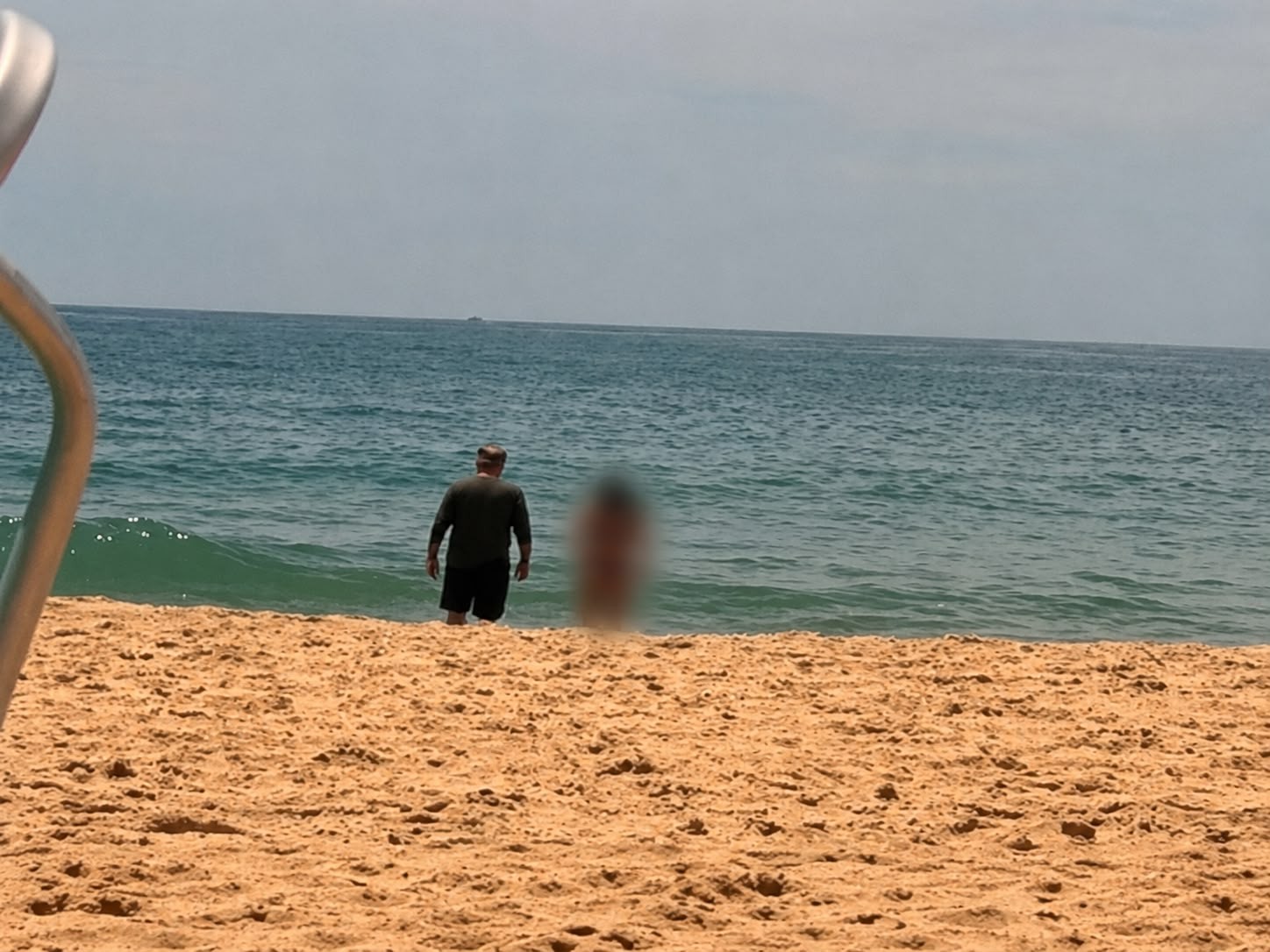 Homem de costas, com camisa escura e bermuda, entra no mar azul-esverdeado de uma praia de areia clara sob céu nublado. Uma pessoa borrada está à direita, e um objeto metálico curvo aparece à esquerda.