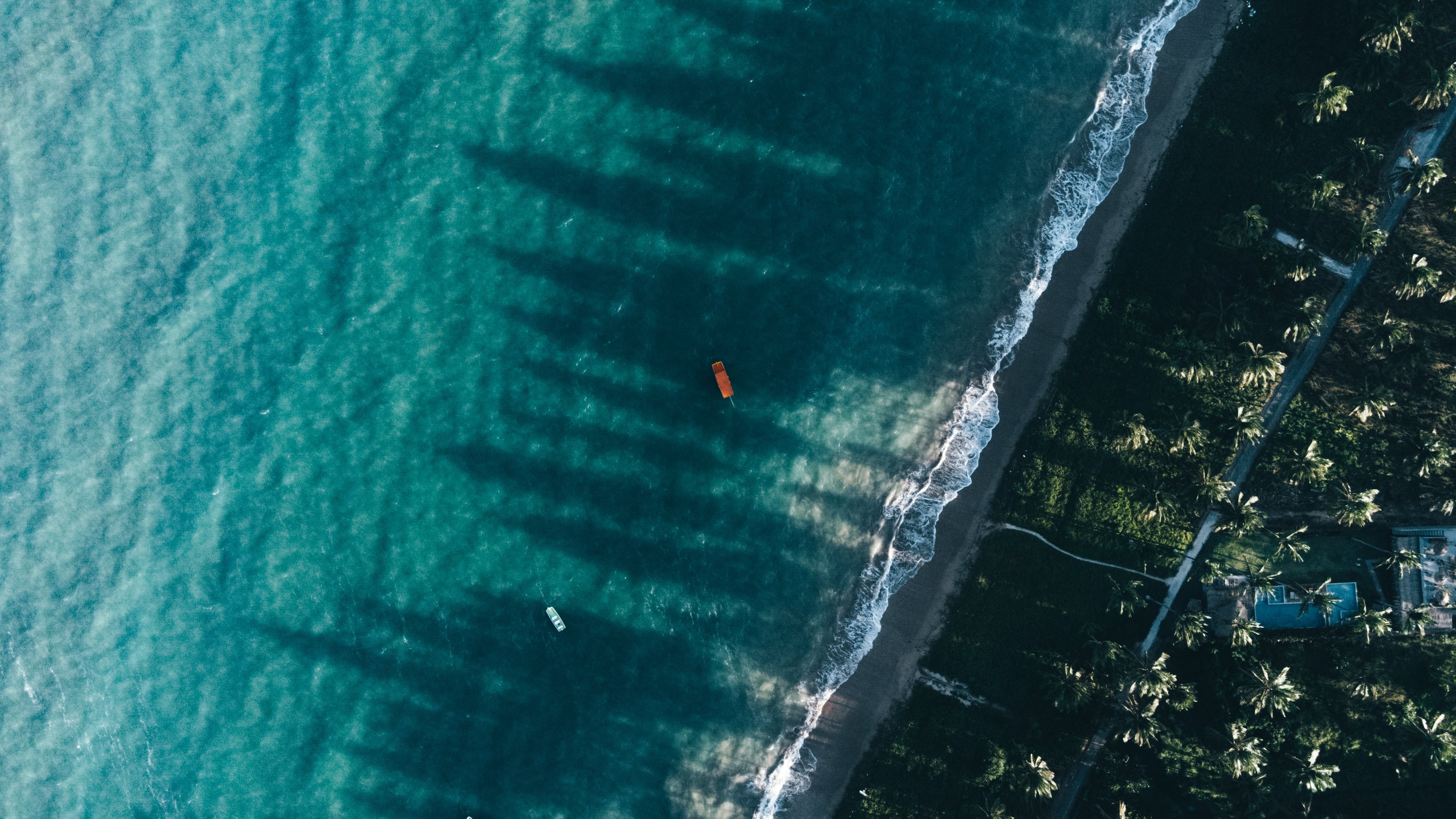 Vista aérea de uma praia tropical com mar azul-turquesa, ondas brancas na areia escura e coqueiros densos. Um barco laranja e outro branco flutuam na água, com sombras alongadas de coqueiros sobre o mar