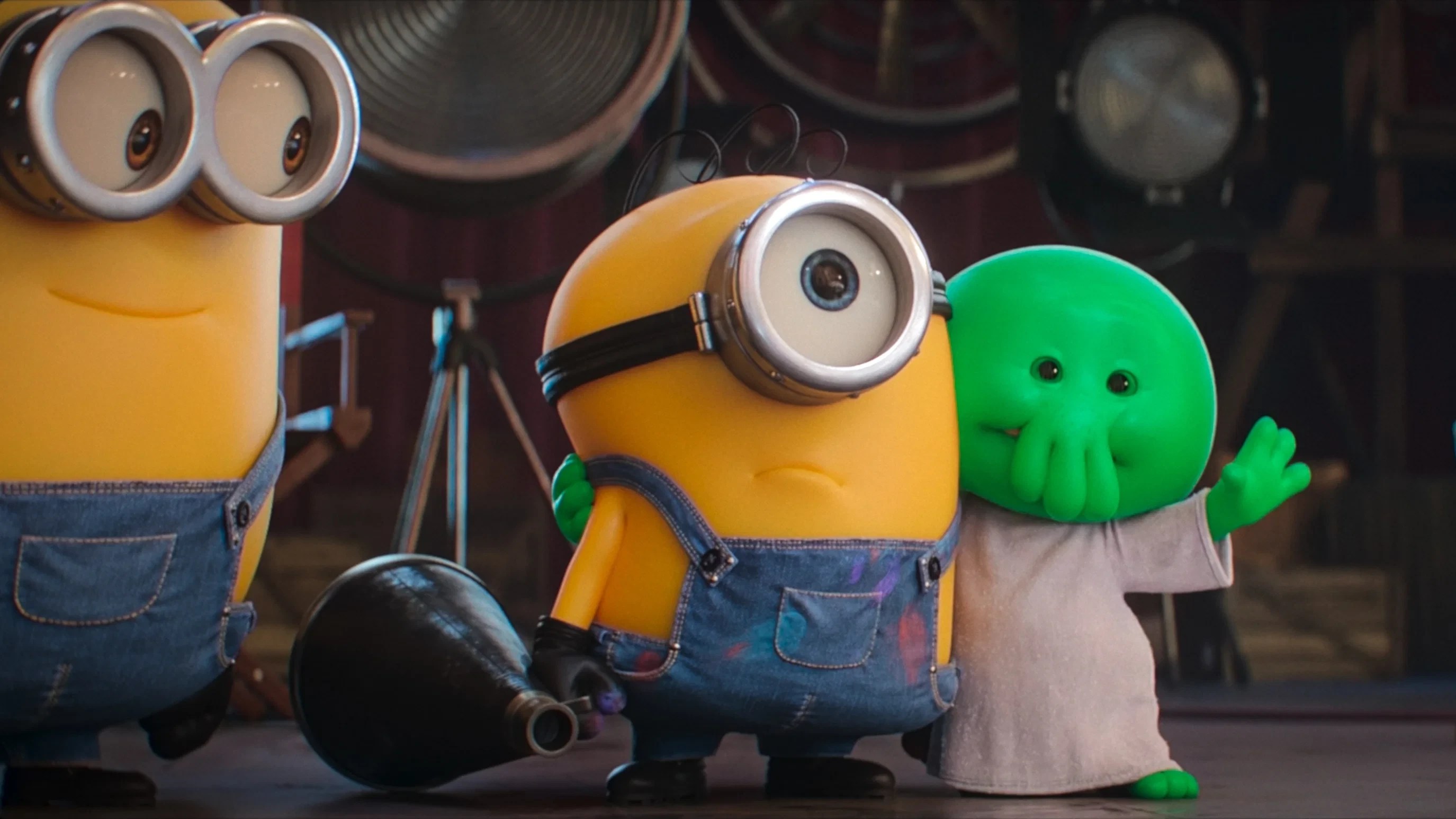 Um Minion amarelo com um olho e óculos prateados, vestindo macacão jeans manchado de tinta, abraça um bebê verde com tentáculos no rosto e roupão bege, que acena. Outro Minion de dois olhos aparece à esquerda, e um megafone preto está no chão. O fundo é escuro com equipamentos de estúdio