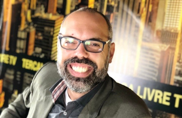 Homem de pele clara, barba e cabelo grisalhos, óculos escuros, sorri amplamente para a câmera. Veste camisa xadrez e jaqueta verde-oliva. Ao fundo, um banner com letras amarelas e pretas, e imagens de prédios