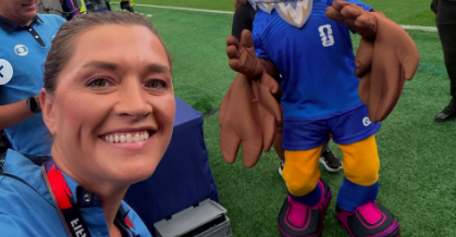 Mulher sorridente de cabelo preso, camisa azul clara e crachá vermelho, tirando uma selfie com um mascote de águia azul, marrom e amarela, em um campo de grama verde